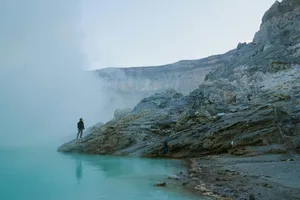 Hiker standing on a foggy ledge beside a turquoise lagoon near jagged cliffs, with distant clouds in the valley