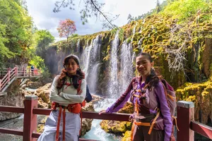 Two trekkers smiling beside a waterfall in Shangri-La, wearing outdoor layers and backpacks on a red-rail viewing platform