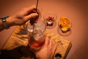 Dalam sesi meracik koktail di Singapura, seorang mixologist menuangkan bahan-bahan dingin ke dalam gelas besar di atas papan kayu.