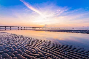 Sandy beach at dusk with footprints leading to a long pier and calm water, with a sky of soft pastel sunset