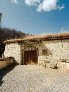 Whitewashed Tibetan homestead doorway at Ferme Liotard in Shangri-La, with carved lintel and tall grass under blue sky