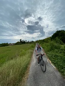 Cyclist pedals along a narrow rural road in Langkawi, past tall grass and hedges under bright, dramatic clouds
