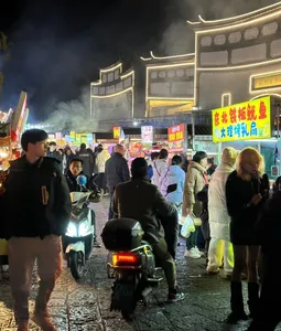 Lijiang night market street crowded with pedestrians and scooters under neon-lit traditional buildings, with misty smoke in the air