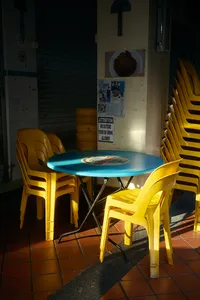 Sunlit café patio table and yellow chairs beside notices on a wall in Eastside Joo Chiat, Singapore, courtyard