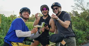 Jackfruit Adventure cyclists in helmets laughing and holding drinks on a bike ride through green paths near Ho Chi Minh City