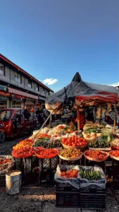 Open-air market in Lijiang Ancient Town piled high with fruit and vegetables in metal trays, under red awnings and blue sky