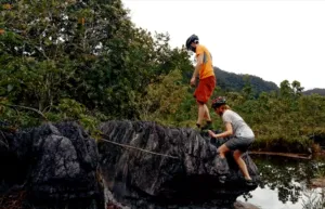 Cyclists climb a dark rock ledge by the river while a fellow rider stands nearby, surrounded by dense jungle near Kuching