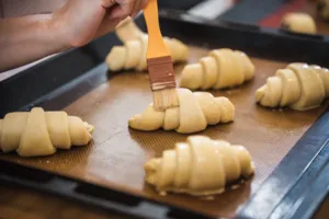 Chef brushing egg wash onto rolled croissant dough on a mesh-lined tray at French Cooking Lab’s Singapore class