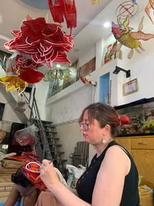 Woman assembling and painting red paper lanterns while other colourful hanging lanterns fill a Ho Chi Minh City craft studio