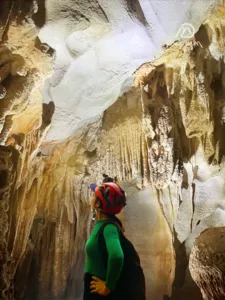 Caver in a green jacket and helmet looks up at illuminated stalactites inside a Phong Nha cave on a Moc Nam Adventure trek