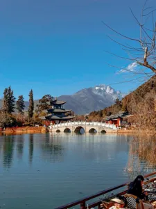 Red arched bridge and traditional pavilion on a calm lake, with snow-capped mountains behind it in Lijiang