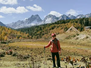Hiker with trekking poles standing in a grassy alpine valley with a small tent as snow-capped mountains loom in Shangri-La