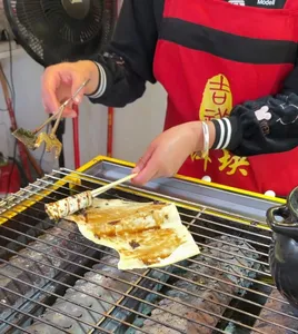 Hands cook thin flatbread on a hot wire rack in Lijiang, with sauce and seasoning at a street food stall