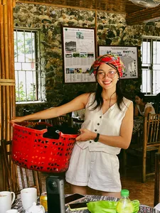 Smiling Ha Giang Expeditions host in a rustic stone room, standing by a table with travel materials and a red basket