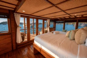 Phinisi cabin bedroom with a neatly made double bed, pillows and sheer curtains, looking out over the water in Labuan Bajo