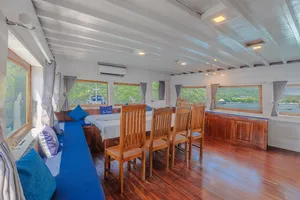 Onboard lounge with blue bench seating, wooden dining chairs and panoramic windows, part of a 3-day join-in Komodo boat trip in Labuan Bajo