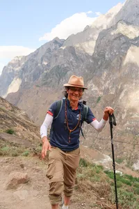 Smiling trekker hiking along a dirt path in Tiger Leaping Gorge, Lijiang, with steep granite peaks towering behind