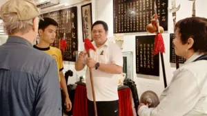 A guide in a white polo explains a lion dance instrument while guests stand around red drapes and calligraphy in Ho Chi Minh City