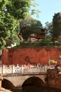Crowds gathered on a stone bridge over water, with a carved red wall and leafy trees in Lijiang Old Town