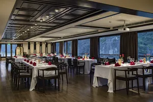 Set dining tables with red napkins and flowers inside a party cruise lounge, looking out over Halong Bay