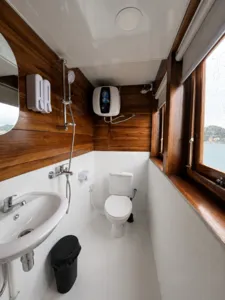 La Famille bathroom interior with a white toilet, wall-mounted sink and water heater, beside a window on a Komodo cruise Labuan Bajo