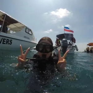 Seorang penyelam di atas bot di perairan Semporna, memakai topeng snorkel dan sut selam, menunjukkan tanda damai sebelum selaman terumbu bersama Bigfin Divers
