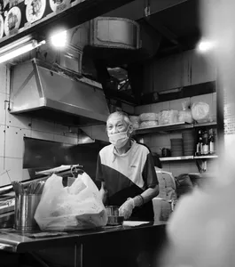 Black-and-white view of an elderly hawker cook preparing food in a Singapore kitchen stall beneath vent hoods and shelves