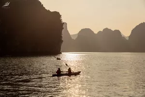 Silhouette of a kayaker paddling across Halong Bay’s calm waters at golden hour between limestone cliffs—Lyra Cruise Collection