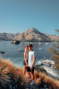 Couple standing on a grassy viewpoint overlooking anchored boats and island hills during a Komodo cruise, Komodo Island