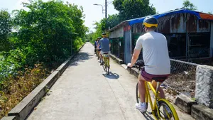 Cyclists pedal along a sunny lane past houses and riverbank vegetation on a Mekong Island ride from Ho Chi Minh City