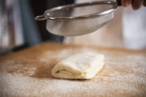 Close-up of croissant dough on a floured worktop as flour is sifted over it in a Singapore kitchen at home
