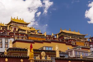 Ornate golden-roofed Tibetan-style palace buildings in Shangri-La against a bright blue sky, with scattered clouds overhead