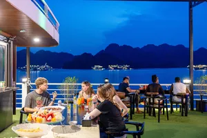 Guests relaxing and dining on the deck of a Halong Bay party cruise at night, with karst silhouettes over the harbour