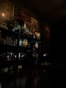 Seorang penghibur membuat isyarat dari belakang kaunter bar di The Magic Bar, Singapura, dengan pencahayaan malap dan botol-botol kaca di sekelilingnya