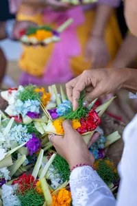 Hands building canang sari leaf baskets with colourful flowers on woven trays during a ceremony in Ubud