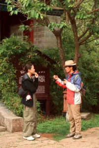 Local guide speaking with travellers beside a stone wall and bamboo garden in Lijiang’s old town courtyard walk