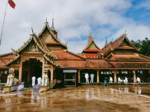 Bangunan kuil yang megah di Xishuangbanna dengan atap berlapis emas, patung, dan halaman yang basah oleh hujan di bawah langit mendung