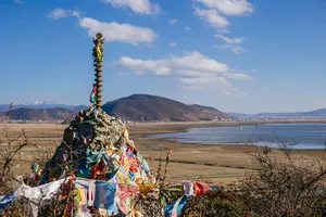 Colourful prayer-flag stone cairn overlooking a wide winter plain and lake in Shangri-La, with low mountains under bright sky