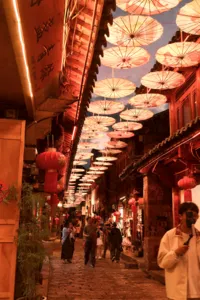 Lantern-covered alley in Lijiang Old Town, warm red light beneath hanging parasols as visitors stroll through the narrow lane