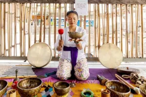 ADDA Yoga sound healer holding a brass singing bowl with resting bowls on a mat, bamboo studio session in Canggu