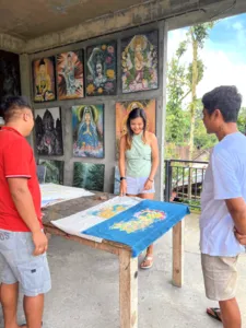 In the Five Arts Studio gallery in Keliki village, a craftsperson and visitors view batik art on a work table in Bali, Ubud