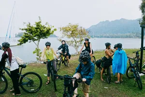 Bicycle Day Langkawi cyclists pause by the bay on Langkawi, standing with bicycles near calm water and distant hills