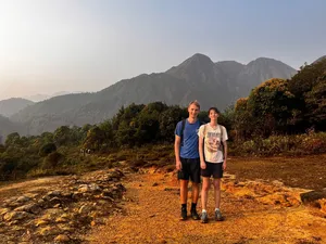 Two travellers walk along a sunlit track through woodland in Sapa, with rugged peaks towering in the distance