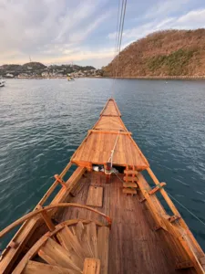 Bow of a wooden phinisi steering through calm water with hills and harbour boats at dusk near Labuan Bajo