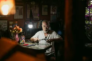 Seorang lelaki membongkok di atas meja yang dipenuhi kertas dan alatan di bawah lampu gantung, untuk kisah sewaan flat Hidden Heritage, permata tersembunyi Singapura