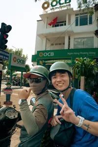 Dua pengendara berhelm di atas skuter vintage di Hoi An, tersenyum dan berpose di samping bendera Vietnam di pusat kota