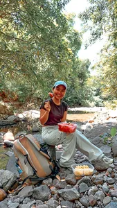 Woman preparing a meal by a rocky stream in the Lombok forest, with bowls and ingredients for the cooking stop