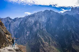 Hiker standing on a rugged cliff path with arms out, looking over steep mountain ranges and valleys in China