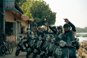 Konvoi tur Vespa di Hoi An dengan para pengendara melambaikan tangan dan tersenyum dari skuter saat menyusuri promenade tepi sungai