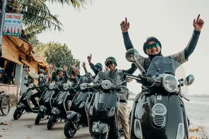 Pengendara skuter berhelm tersenyum dan melambaikan tangan ke kamera saat mengikuti perjalanan Hoi An Backroad Tours dengan Vespa bergaya vintage